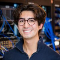 Young man with brown hair and round glasses smiling in a tech workspace with blue cables in the background.
